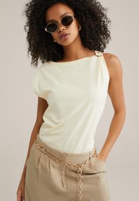 Cream-colored sleeveless top with an asymmetrical neckline and gold hardware accent, paired with beige high-waisted pants featuring a chain belt.