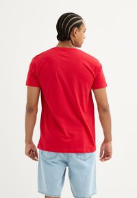 Camiseta de algodón roja con mangas cortas y cuello redondo. Textura suave, diseño sencillo, combinada con pantalones cortos de mezclilla azul claro.