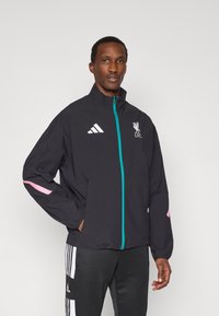 Μαύρο αδιάβροχο μπουφάν με μεταλλικό τσάι φερμουάρ σε τιρκουάζ απόχρωση, ροζ λεπτομέρειες στα μανίκια και λογότυπο της Λίβερπουλ FC στο στήθος, κατασκευασμένο από ελαφρύ ύφασμα.