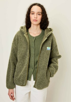 Fleece jacket in olive green with a zip front and hood. Features a fluffy texture and patch detail, paired with a matching sweater underneath.
