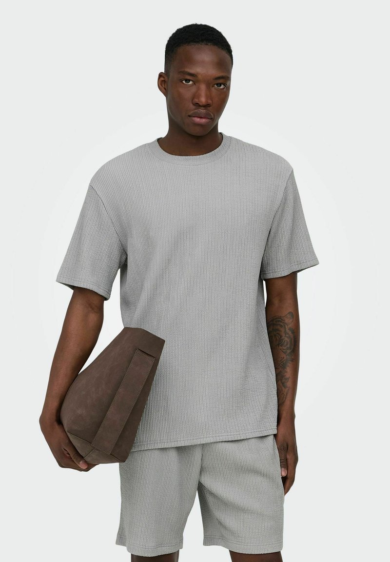 Homme portant une chemise texturée gris clair à manches courtes et un short, tenant un grand sac rectangulaire marron dans sa main gauche.