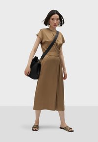 Brą dress w kolorze brązowym, z krótkim rękawem, uszyta z miękkiego, gładkiego materiału, o fasonie kopertowym. Czarna torba typu bucket o teksturowanym wykończeniu i cienkim pasku. Płaskie sandały w paski.