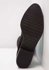 Botín de ante negro con suela de goma texturizada, que presenta un patrón de zigzag y un logotipo en relieve. Talla marcada como 37.