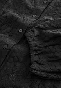 Schwarze Bluse aus Stoff mit floralen Mustern, ausgestattet mit runden schwarzen Knöpfen und strukturierten Rüschen. Filigrane Spitzenverzierungen sorgen für ein detailreiches Design.