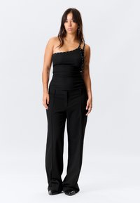 Μαύρο τοπ με έναν ώμο και διακοσμητικά καρφιά στο λουρί, συνδυασμένο με μαύρο παντελόνι φαρδιάς γραμμής. Ομαλή υφή υφάσματος και εφαρμοστή γραμμή.