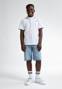 Vit pikétröja med marinblå krage, ljusblå denimshorts, vita sneakers och svartrandiga strumpor. Leende barn som står.