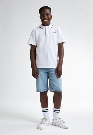 Weißes Polohemd mit einem marineblauen Kragen, hellblaue Jeansshorts, weiße Turnschuhe und schwarzgestreifte Socken. Lächelndes Kind steht.