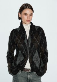 Cardigan a maglia con motivo argyle in nero e grigio, con zip davanti, polsini a costine e materiale in lana testurizzata, indossato sopra un dolcevita beige.