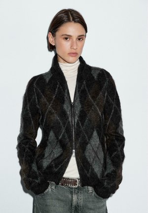 Cardigan a maglia con motivo argyle in nero e grigio, con zip davanti, polsini a costine e materiale in lana testurizzata, indossato sopra un dolcevita beige.