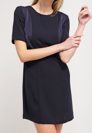 Femme portant une robe noire à manches courtes avec des panneaux latéraux en satin, debout, les bras croisés et une main touchant le poignet opposé.