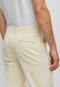 Pantaloni color crema con una texture liscia, dotati di una vita standard, taglio dritto e due tasche posteriori. Piccola etichetta con logo visibile.