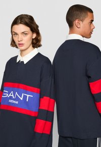 Marineblå langermet polo med røde og blå striper, med en hvit krage og "GANT SPORTSWEAR" logo på fronten.