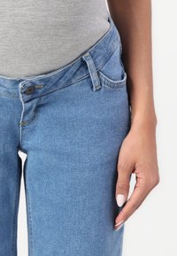 Ženská ruka odpočívající u boků, v světle modrých džínách a šedém přiléhavém topu, na jednorázovém bílém pozadí.