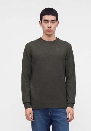 Jeune homme aux cheveux courts noirs portant un pull vert foncé à col rond et un jean bleu, debout devant un fond clair uni.