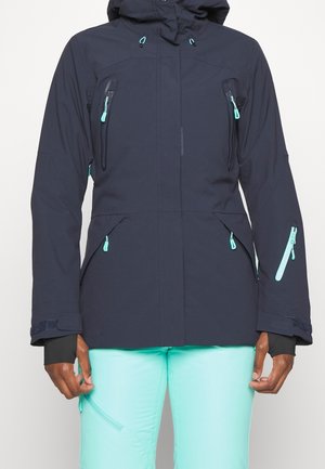 Persoon die een donkerblauwe capuchonjas draagt met teal ritsen en licht teal broek, staand tegen een effen achtergrond.