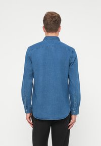 Džínsová košeľa v tmavomodrej farbe, s golierom na gombíky, dlhými rukávmi s gombíkovými manžetami a zakriveným spodným okrajom. Hladká textúra látky.