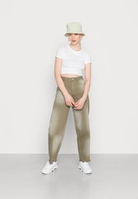 Camiseta branca cropped, calças de cintura alta verde-oliva com uma textura desbotada e sapatilhas brancas; acessorizado com um chapéu de balde em tom claro.