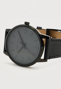 Calvin Klein schwarze Uhr mit einem strukturierten grauen Ziffernblatt, schlanken schwarzen Zeigern und einem schwarzen Lederarmband mit subtilen Stickdetails.