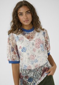 Junge Frau mit lockigem Haar, die eine durchsichtige Bluse aus floraler Spitze mit blauen Akzenten und einer weißen Unterlage trägt und mit der Hand in der Tasche posiert.