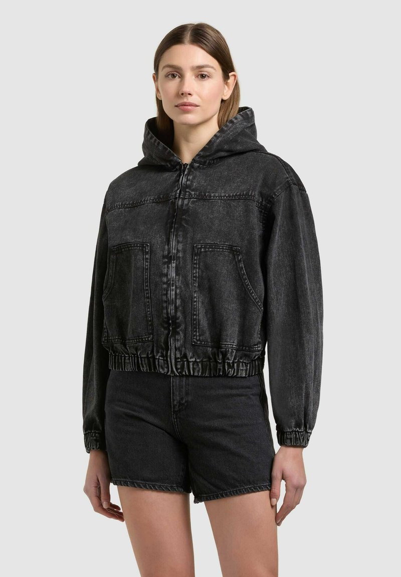 Frau trägt schwarze, verwaschene Denim-Jacke mit Reißverschluss und Kapuze sowie passende Shorts, steht vor einfachem Hintergrund.