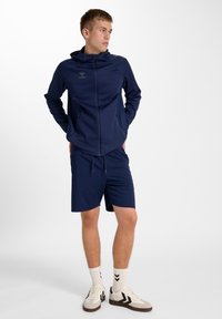 Marineblaues Sportoutfit mit einer Jacke zum Reißverschluss und Kapuze sowie Shorts. Die Jacke hat zwei Reißverschlusstaschen und besteht aus glattem Stoff.