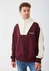 Felpa in pile color mogano e crema con colletto alto, zip a metà e taglio quadrato. Presenta un ricamo del logo sul davanti.
