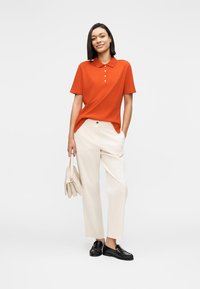 Πορτοκαλί πόλο πουκάμισο με γιακά και λεπτομέρειες με κουμπιά, συνδυασμένο με εκρού παντελόνι φαρδύ στο πόδι και μαύρα μοκασίνια· κρατώντας μια μπεζ τσάντα χειρός.