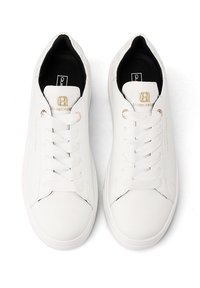 Zapatillas blancas con un exterior de cuero suave, punta redonda, suela de goma plana y cierre con cordones. Presenta acentos del logo en dorado en la lengüeta.