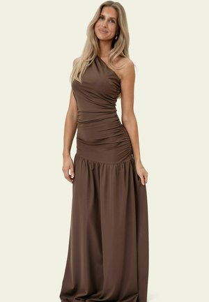 Frau mit langen blonden Haaren, die ein ärmelloses, gerafftes, dunkelbraunes Ein-Schulter-Kleid trägt und vor einem schlichten hellen Hintergrund steht.