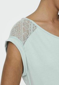 Jasnoniebieska koszulka z luźnym krojem, koronkowym wykończeniem na ramieniu i okrągłym dekoltem. Wykonana z miękkiej bawełny, z krótkimi rękawami.