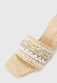 Beige vevd mule-sandal med flat, firkantet tå, utsmykket stropp med dekorative gull-, sølv- og perleaksenter. Glatt innvendig fôr.