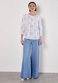 Vit blus med blå blommönster, lös passform och trekvartslängd på ärmarna, i kombination med vida ljusblå jeans och vita klacksandaler.