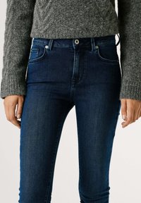 Vaqueros ajustados azul oscuro con diseño texturizado, estilo de cinco bolsillos y cierre con botón metálico, combinados con un suéter gris de punto.