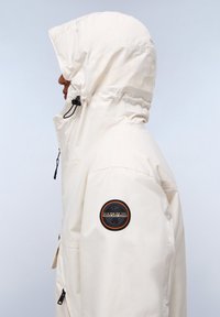 Lichtcrème kleurige parka met een capuchon, voorzien van een zwarte patch op de linkermouw en een verstelbare trekkoord bij de hals. Soepele textuur.