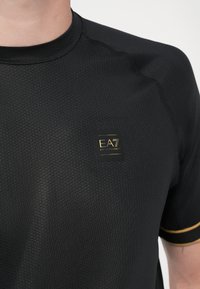 Čierne atletické tričko s textúrovaným vzorom, okrúhlym výstrihom a zlatým akcentom na rukávoch. Obsahuje malé logo EA7 na hrudi.