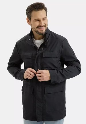 Mężczyzna z brodą i ułożonymi włosami uśmiecha się, zapinając czarną kurtkę z czterema przednimi kieszeniami na białą koszulę.