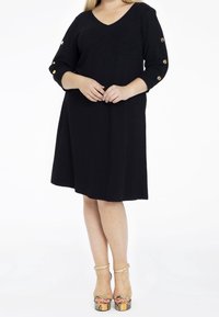 Robe noire avec un décolleté en V et des manches trois quarts, ornée de boutons ronds en or. Silhouette évasée avec un tissu texturé.