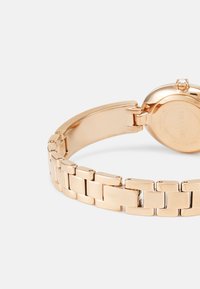 Reloj dorado con pulsera de eslabones de metal y caja redonda. Cuenta con un pequeño cristal en la corona y un logotipo en el cierre.