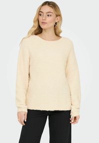 Cremefarbener flauschiger Pullover mit rundem Ausschnitt, langen Ärmeln und einer leicht lockeren Passform, getragen über schwarzen Hosen.