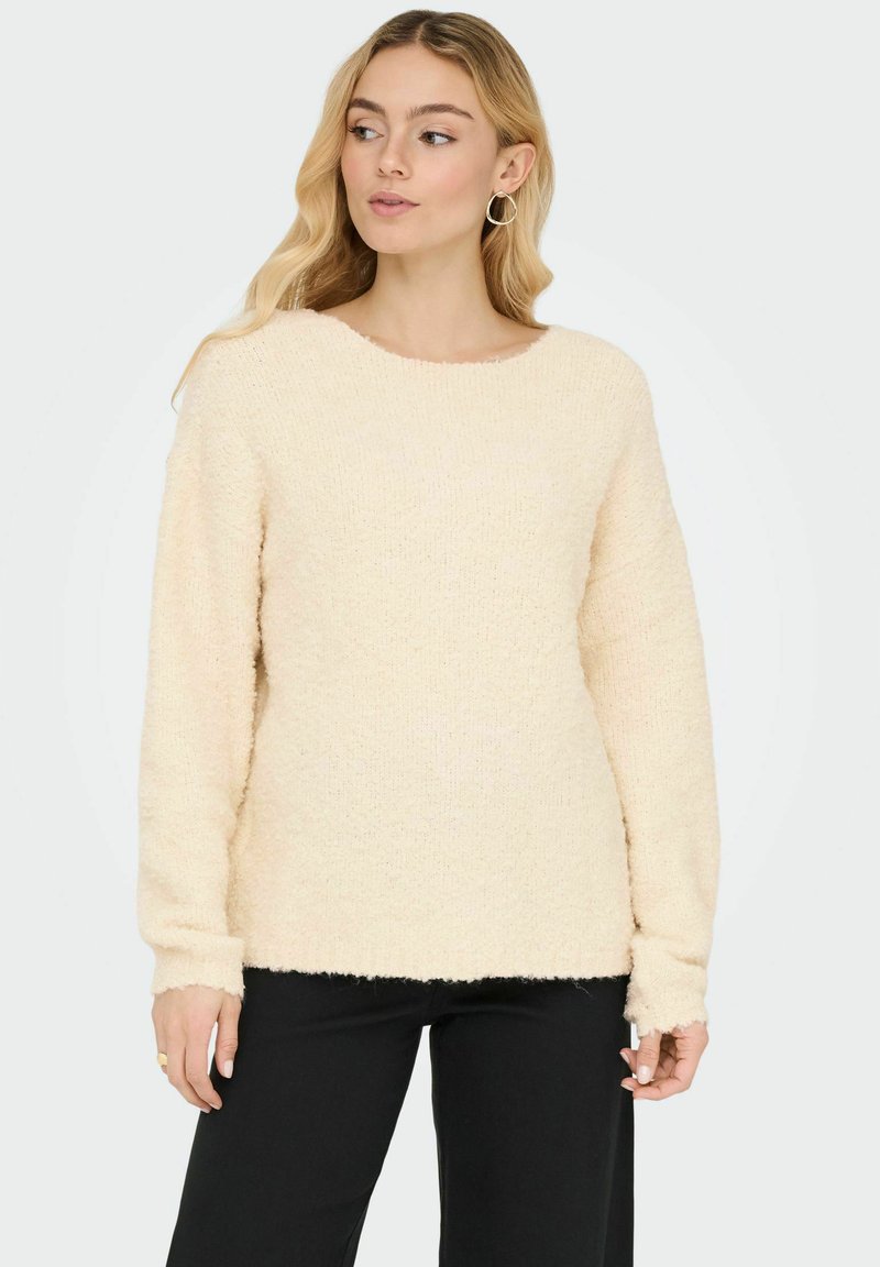 Cremefarbener flauschiger Pullover mit rundem Ausschnitt, langen Ärmeln und einer leicht lockeren Passform, getragen über schwarzen Hosen.