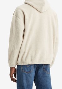 Felpa con cappuccio in pile beige con superficie testurizzata, cappuccio ampio e spalle scivolate, abbinata a jeans di denim blu con tasche posteriori.