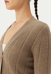 Ljusbrun kofta gjord av ribbat tyg med djup V-ringning och texturerad detaljering; har långa ärmar och en avslappnad passform.
