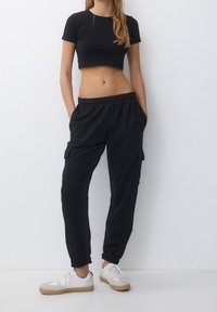 T-shirt noir court à manches courtes, associé à un pantalon cargo noir avec poches latérales et des poignets élastiques, porté avec des baskets blanches.