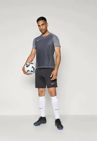 Grå treningsskjorte med korte ermer og hvite detaljer, svarte shorts med oransje logo, hvite sokker og svarte fotballsko som holder en fotball.