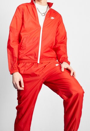 Osoba ubrana w czerwony dres Nike z białym zamkiem i logotypami, białą koszulkę oraz srebrny naszyjnik z łańcuszkiem na jednolitym tle.
