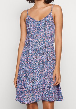Femme portant une robe bleue à fleurs sans manches avec des bretelles fines et une ceinture nouée, debout devant un fond uni.
