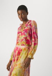 Blusa floreale in rosa, giallo e verde, con scollatura a V arricciata e maniche lunghe, abbinata a una gonna plissettata. Tessuto leggero e trasparente.