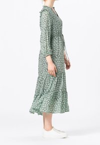 Robe fleurie verte avec un col à volants, des manches trois-quarts et une jupe à volants. Portée avec des baskets blanches. Tissu léger et fluide.