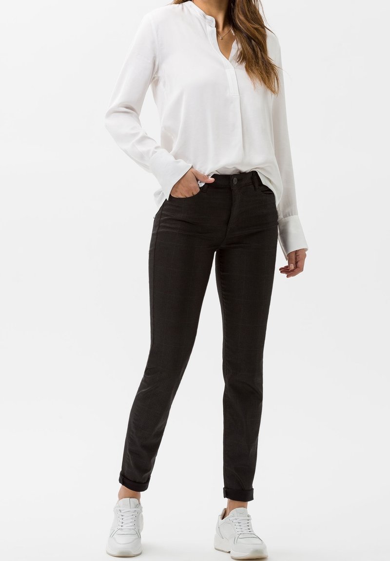 Pantalones negros con patrón y corte ajustado, que cuentan con un cierre de botón, bolsillos laterales y puños enrollados por encima del tobillo, combinados con una blusa blanca.