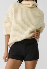 Pull en maille côtelée crème avec un col roulé, associé à un short noir sur mesure. Le tissu semble doux et texturé.